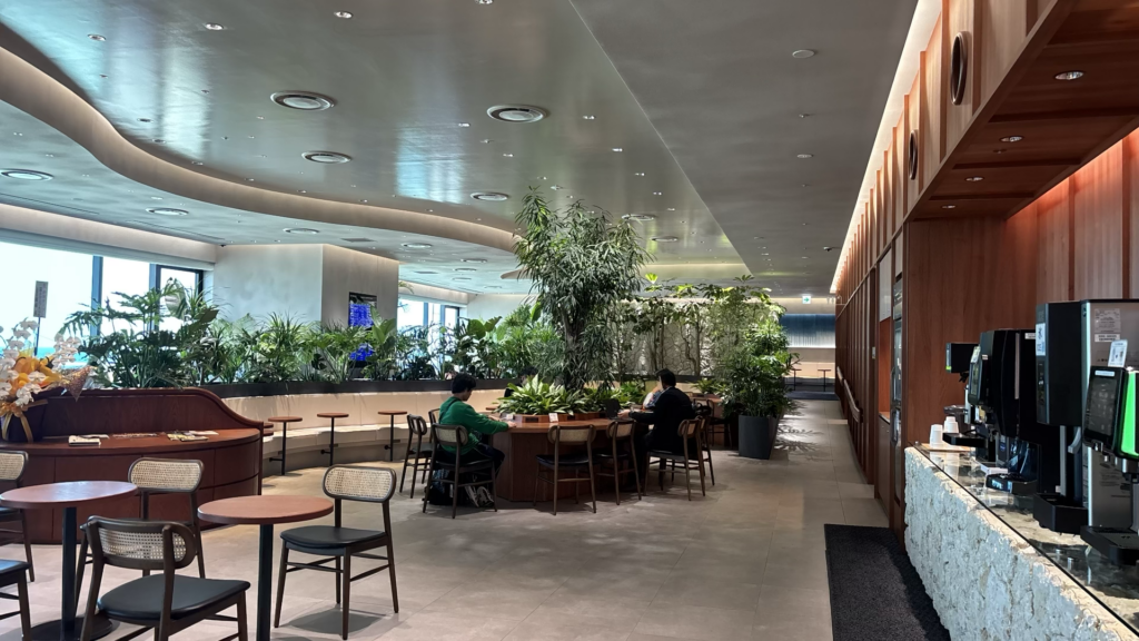 植物に囲まれた明るいカフェラウンジの店内。テーブル席で過ごす人々と、右側にコーヒーマシンが並ぶ。